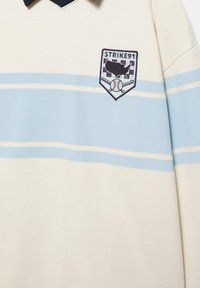 Krémová a svetlomodrá pruhovaná košeľa s námorníckym golierom a nášivkou „STRIKE91“ na hrudi, ktorá zobrazuje baseball, baseballové pálky a mapu USA.