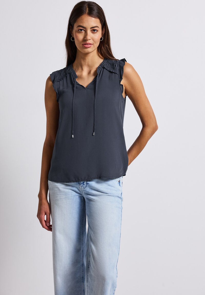 Haut sans manches gris foncé avec un décolleté en V, des détails froncés sur les épaules et des accents à cordon. Tissu lisse et léger, associé à un jean bleu clair.