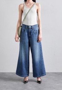 Pantalones vaqueros de corte ancho de mezclilla azul con un lavado claro, cinco bolsillos y un cierre clásico de botón, combinados con una camiseta sin mangas blanca y tacones negros.