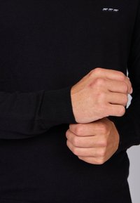 Pull en tricot noir avec des poignets côtelés ; présente un petit logo d'animal blanc sur la poitrine. Texture lisse et coupe classique.