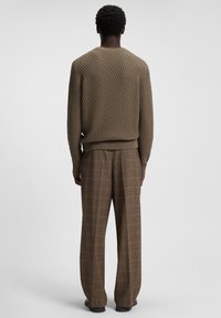Mann, der mit dem Rücken zur Kamera steht und einen strukturierten braunen Pullover sowie weite braune Karohosen trägt, vor einem einfarbigen Hintergrund.
