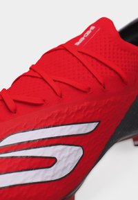 Κοντινό πλάνο ενός κόκκινου αθλητικού παπουτσιού με υφή στην επιφάνεια, κόκκινες κορδόνια και μεγάλο λευκό στυλιζαρισμένο λογότυπο "S" στο πλάι.