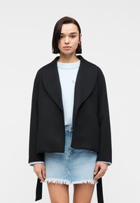 Jeune femme aux cheveux lisses et foncés portant une veste noire ouverte, un pull bleu clair et une mini-jupe en denim clair effilochée, sur fond blanc.