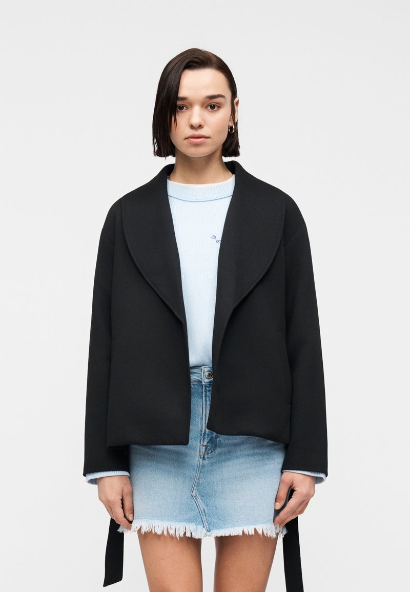 Jeune femme aux cheveux lisses et foncés portant une veste noire ouverte, un pull bleu clair et une mini-jupe en denim clair effilochée, sur fond blanc.