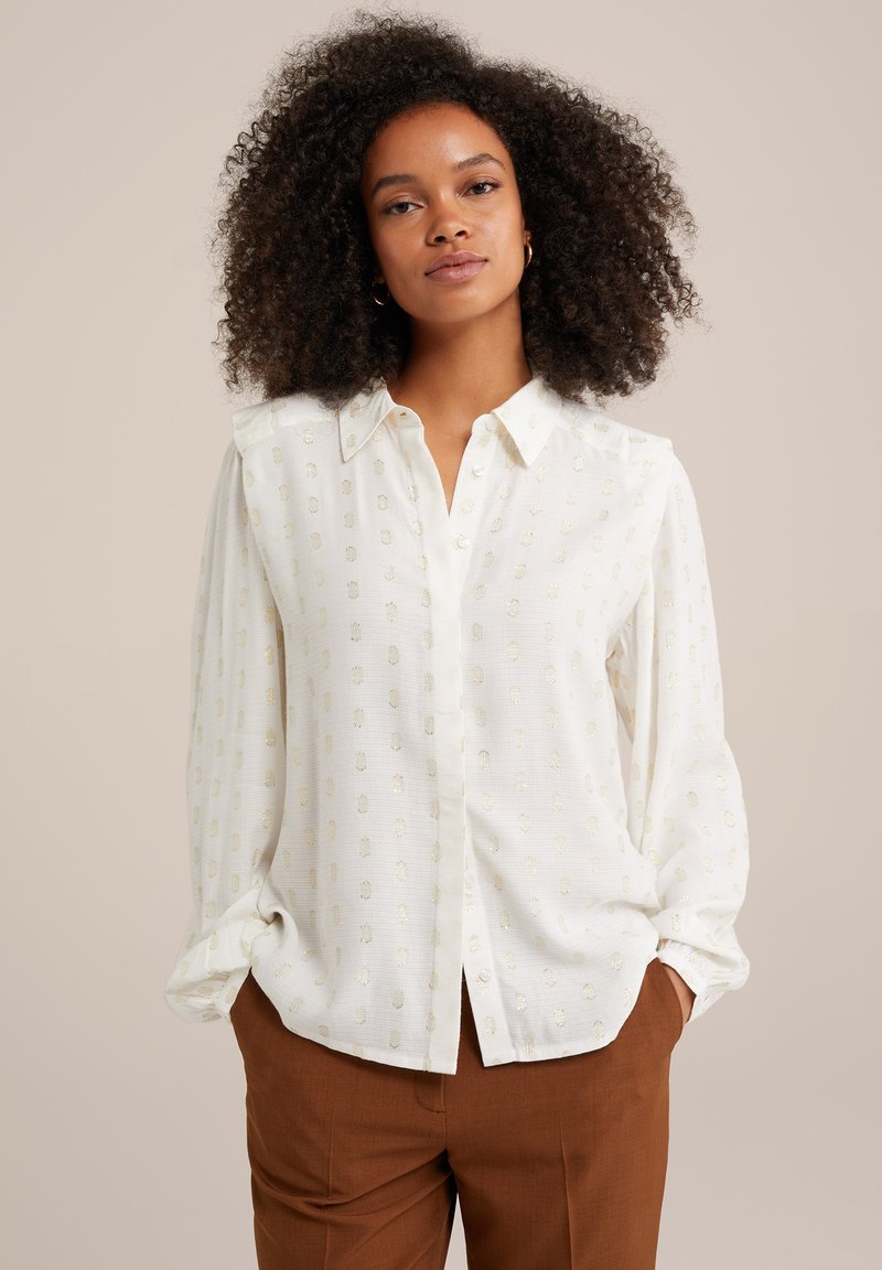 Camicia bianca con bottoni, caratterizzata da un sottile motivo testurizzato e dettagli metallici. Presenta un colletto, maniche lunghe e una vestibilità rilassata. Abbinata a pantaloni marroni.