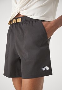 Svarta träningsshorts med elastisk midja och tan-spänne, med sidofickor och en vit logotyp på nedre vänstra sidan.