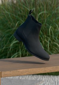 Botte cheville en cuir noir au design minimaliste, avec des panneaux latéraux en élasthanne et une texture lisse et mate, posée sur une surface en bois.