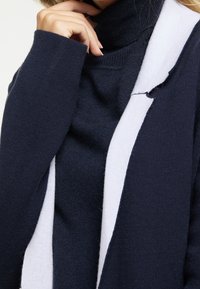 Marineblauer Strickcardigan mit weißem Akzent an den Kanten, mit langen Ärmeln und einem Rollkragenpullover darunter, der weiche Texturen zeigt.