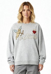 Femme portant un sweat-shirt gris avec un cupidon tenant un arc et des flèches, un cœur rouge, et les mots "Je T'aime" en écriture cursive.