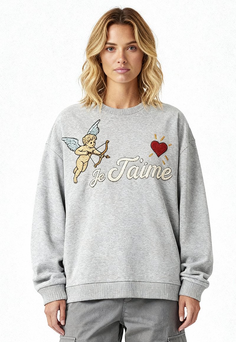 Femme portant un sweat-shirt gris avec un cupidon tenant un arc et des flèches, un cœur rouge, et les mots "Je T'aime" en écriture cursive.