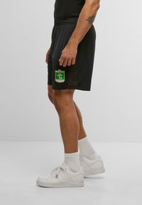 Man wearing black sports shorts with "Fussballclub Kitzbühel" logo, white socks, and white Nike Air sneakers standing on gray floor.