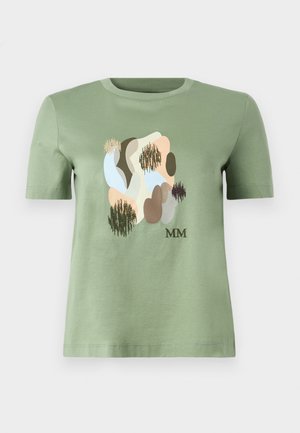 Frau trägt grünes abstraktes Print-T-Shirt und beige weite Hosen, steht vor weißem Hintergrund mit einer Hand in der Tasche.