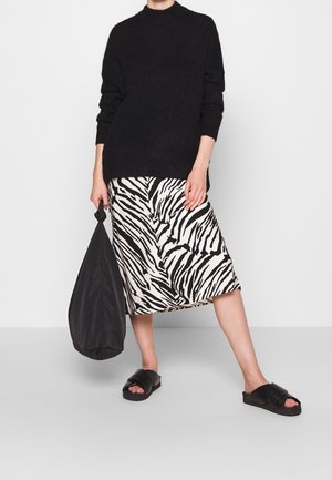 Personne portant un pull noir, une jupe imprimée zèbre noir et blanc, des sandales slides noires, tenant un grand sac noir sur un fond uni.