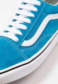 Modrá semišová teniska s bílými tkaničkami a kontrastním bílým proužkem po straně. Má pryžovou podešev s černým akcentem kolem spodní části.