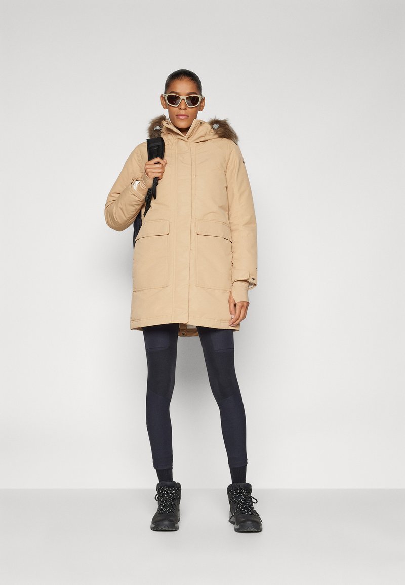 Parka beige avec capuche en fourrure, deux poches avant et fermeture éclair. Le modèle porte des leggings noirs et des bottes noires robustes. Des lunettes de soleil complètent le look.