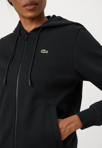Juodas zip-up gobtuvu džemperis, pagamintas iš tekstūruotos medžiagos, su mažuoju žaliu siuvinėtu logotipu krūtinėje ir reguliuojamu raišteliu gobtuve.