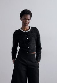 Schwarze, flauschige Strickjacke mit einem mit Juwelen besetzten Kragen, runden Knöpfen und langen Ärmeln, kombiniert mit hochgeschnittenen schwarzen Hosen.