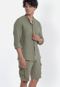 Camicia in lino verde oliva con colletto a banda, maniche lunghe e chiusura con bottoni, abbinata a pantaloni cargo coordinati con tasche laterali.