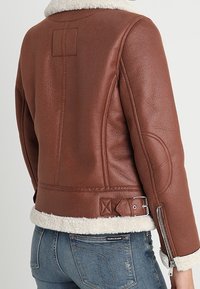 Giacca in pelle marrone con colletto in pelliccia, zip laterale e dettagli testurizzati. Caratterizzata da un design aderente e accenti regolabili in vita.