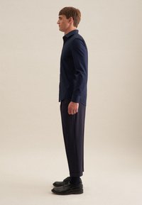 Man die een donkerblauw overhemd met knopen draagt, donkere broek en zwarte nette schoenen, staand in profiel tegen een eenvoudige neutrale achtergrond.