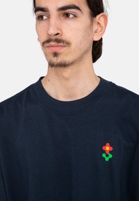 Camiseta de algodón azul marino con cuello redondo y mangas cortas. Presenta un logo multicolor (rojo, amarillo, verde) en el lado izquierdo del pecho.
