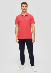 Czerwony golf polo z kołnierzykiem i krótkim rękawem, z białymi detalami i logo. Noszony z ciemnymi spodniami i białymi trampkami, stojący prosto.