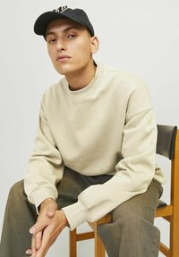 Sudadera beige con cuello y puños acanalados, combinada con pantalones oscuros de estampado, sentado en una silla de madera frente a un fondo claro.