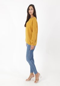 Musztardowo-żółty sweter na drutach o fakturowanym wzorze, zestawiony z dopasowanymi niebieskimi dżinsami i przezroczystymi sandałami na obcasie, ustawiony nieco bokiem.