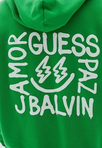 Sudadera verde con texto gráfico blanco "Major Guess Paz J Balvin" y una cara sonriente con ojos de rayo en la parte trasera.