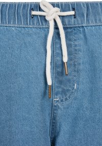 Gros plan sur un short en denim bleu clair avec une ceinture élastique et un cordon blanc noué avec des embouts métalliques.