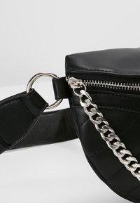 Coin de sac en cuir noir avec une fermeture éclair argentée, une chaîne argentée attachée et une lanière noire reliée par un anneau argenté.