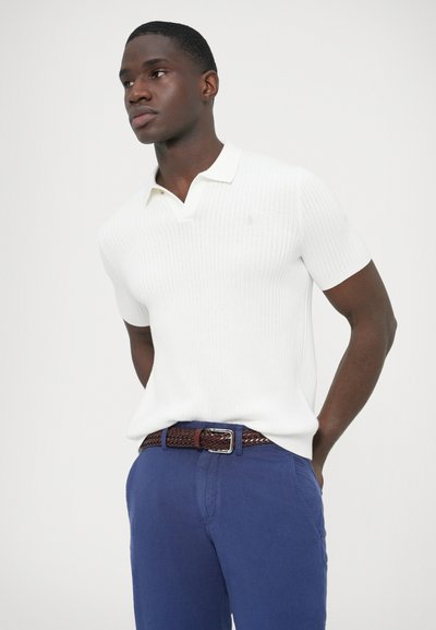 Biela rebrovaná polokošeľa s krátkymi rukávmi, v kombinácii s modrými nohavicami a hnedom pleteným opaskom, s klasickým golierom a decentným logom.