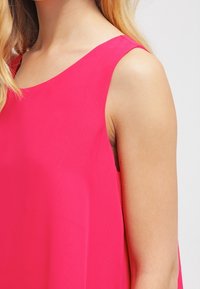 Kobieta w jasnoróżowej bezrękawnikowej bluzce, z widocznymi blond włosami na jasnym tle.