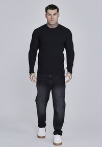 Mustad mustade pikkade varrukatega henley särk ribatud tekstuuriga, nööbiga kaeluse ning reljeefse logoga, ühendatud tumedate teksade ja valgete tossudega.