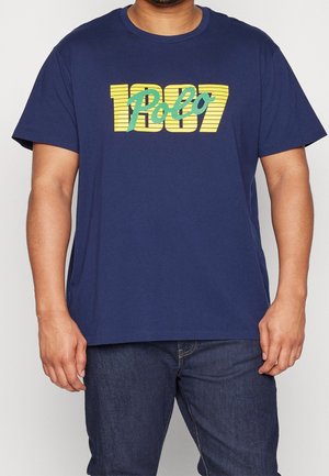 Homme portant un t-shirt bleu marine avec le texte jaune et vert « 1987 Polo » imprimé sur le devant, associé à un jean bleu foncé.