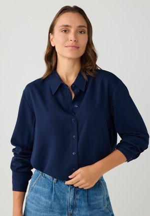Camicia button-up blu navy realizzata in un tessuto morbido, dotata di colletto, maniche lunghe e vestibilità rilassata. Presenta una parte anteriore piatta e una leggera lucentezza.