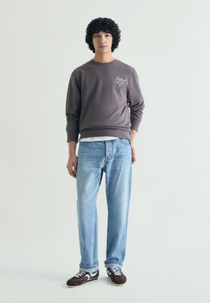 Joven con cabello rizado que lleva una sudadera oscura con texto blanco, vaqueros azul claro y zapatillas oscuras sobre un fondo claro y liso.