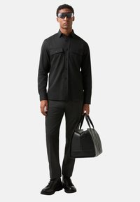 Black button-up shirt with chest pockets, dark grey trousers, black leather bag, and black boots. Model wears sunglasses. Minimalist design.