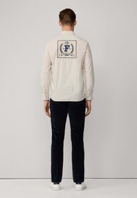 Lichtbeige langetermijnhemd met een marineblauw gedrukt logo en laurierontwerp op de achterkant. Gecombineerd met donkere broek en witte schoenen.