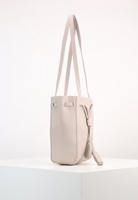 Beżowa skórzana torebka w kształcie wiadra z dwoma długimi paskami, płaskim dnem, bocznymi frędzlami i minimalistycznym designem. Gładka tekstura, pionowe przeszycia.
