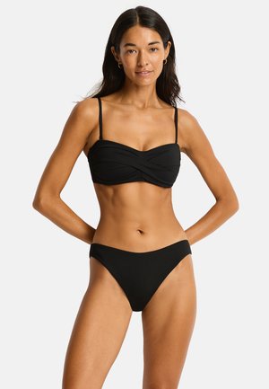 Femme aux longs cheveux foncés portant un bikini noir avec un haut bandeau torsadé et un bas taille haute, debout les mains derrière le dos.
