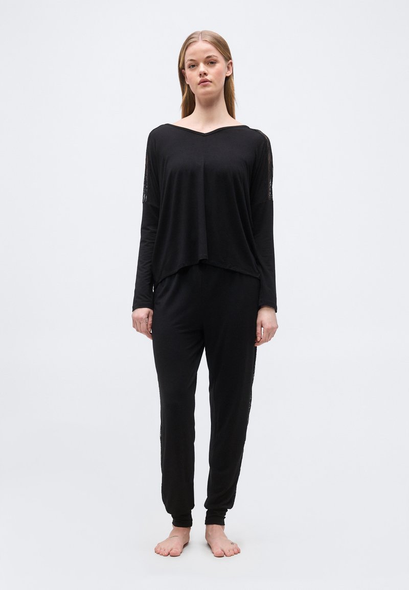 Jeune femme debout pieds nus, portant un haut ample à manches longues noir et un pantalon noir assorti, sur un fond clair uni.