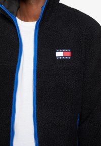 Czarna polarowa kurtka z niebieskim zamkiem błyskawicznym, z kwadratowym logo "TOMMY JEANS" na klatce piersiowej. Miękka tekstura, pełny zamek z przodu, casualowy design.