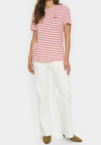 Mujer vestida con una camiseta de rayas rojas y blancas de mangas cortas, pantalones anchos blancos y tacones marrones de punta en un fondo blanco.