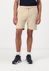 Pantalones cortos de algodón beige con cintura elástica y cordón. Cuenta con bolsillos laterales y un logo en el dobladillo. Combinados con zapatillas deportivas azul marino.