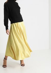 Femme portant un pull en maille noir, une jupe fluide en satin jaune et des sandales à talons hauts noires, debout devant un fond blanc uni.