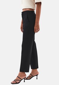 Persona vestida con pantalones cortos de tiro alto negros y bolsillos laterales, junto con sandalias de tacón alto negras con tiras, de pie de lado.