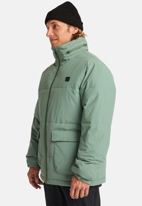 Giacca verde e morbida con colletto alto, zip frontale e due tasche laterali. Presenta un'etichetta con logo e una texture liscia. Indossata con un berretto nero.
