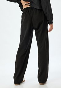 Schwarze weite Hosen mit glatter Textur, elastischem Bund und lockerer Passform, kombiniert mit hellen Schuhen.