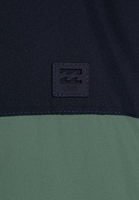 Navy and green color-blocked fabric, featuring a textured surface and a square black logo patch with wave design and "A/DIV." text.